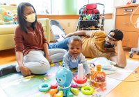 A baby boy surrounded by his smiling mom and dad sits on the floor of his hospital room surrounded by toys