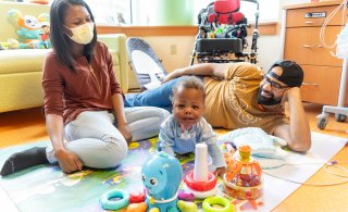 A baby boy surrounded by his smiling mom and dad sits on the floor of his hospital room surrounded by toys