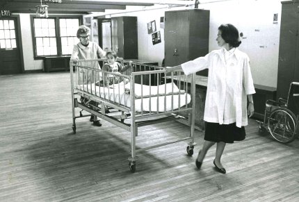 Small child being transported by two nurses in a rolling crib